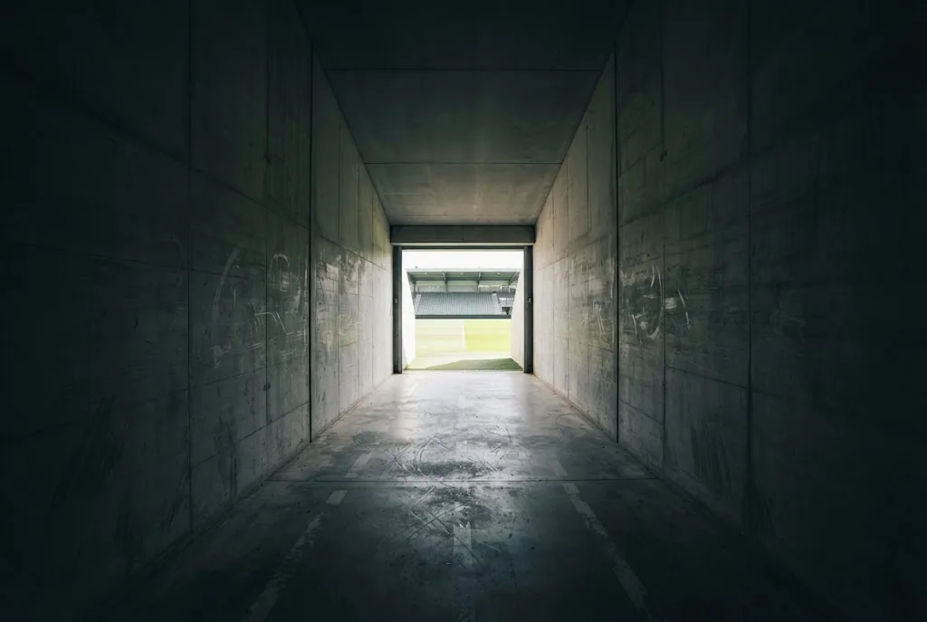 A quiet stadium tunnel leading out towards daylight and the edge of a pitch